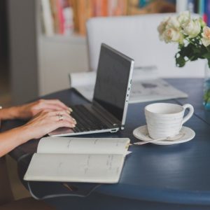 Händer som skriver på en laptop, en uppslagen bok och en kopp kaffe illustrerar flitiga händer som du kan hyra in via en redaktörstjänst.